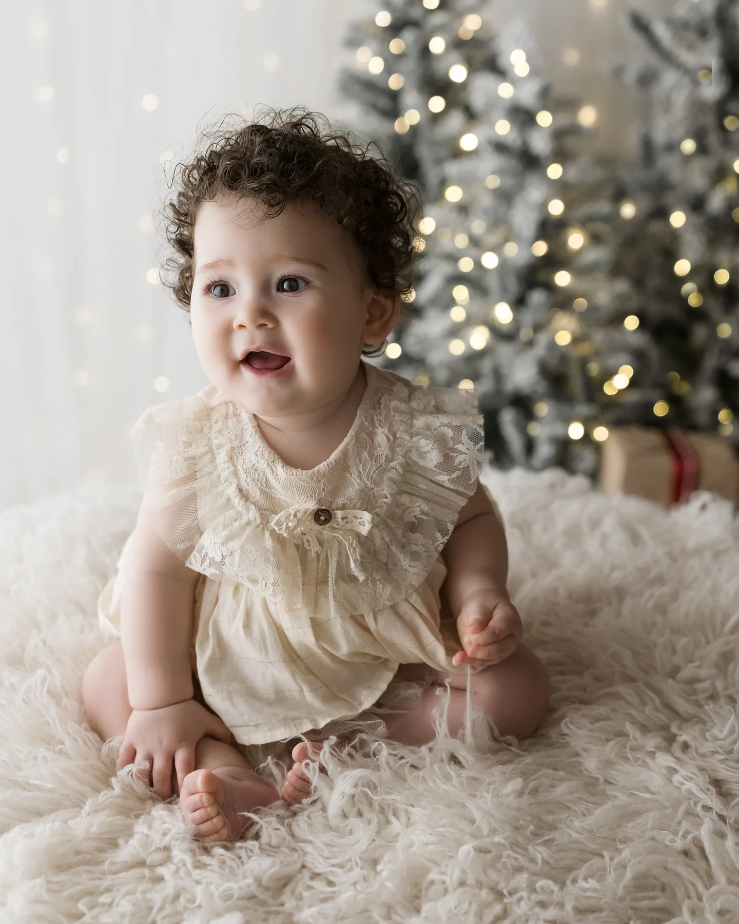 Bambina riccia con il vestito color panna seduta sulla pelliccia davanti all&rsquo;albero